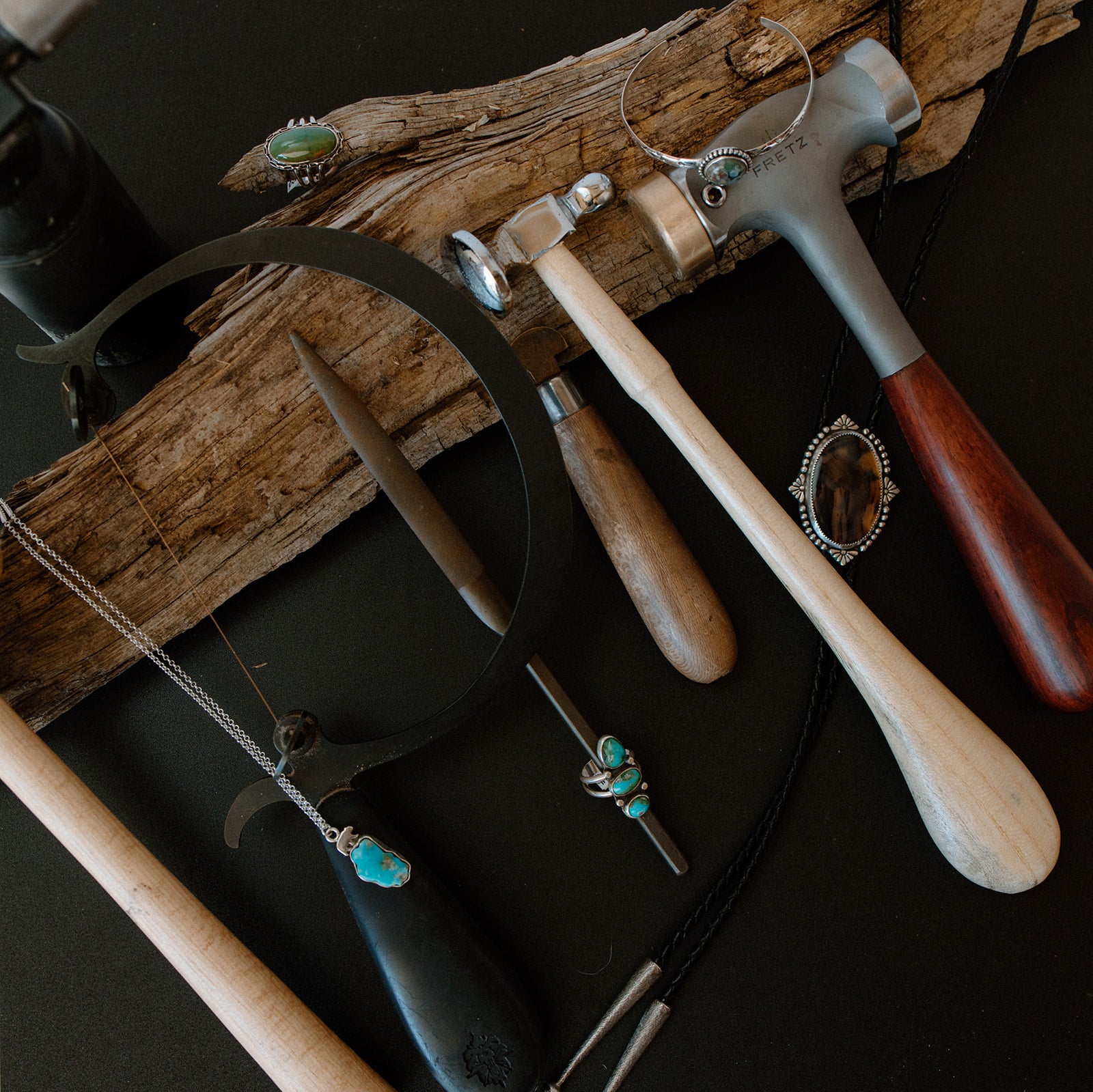 A styled flat-lay of jewelry-making tools and finished jewelry pieces arranged on a dark surface with a piece of driftwood. Visible items include a silver ring mandrel with turquoise and brown stone settings, a hammer with a wood handle, a metal mallet, and several silver necklaces with turquoise and brown stone pendants.